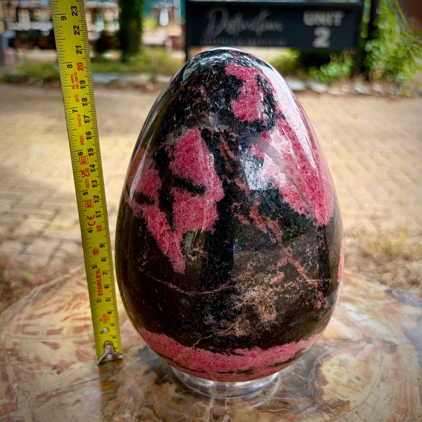 A polished rhodonite crystal egg resting on a clear stand next to a yellow measuring tape.